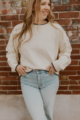 Woman wearing a beige sweater and light blue jeans standing against a brick wall.